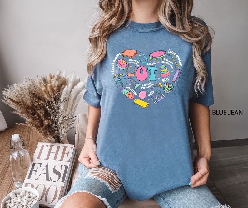May include: A blue t-shirt with a heart-shaped design featuring various colorful illustrations of toys and the text "visual motor", "fine motor", "social", "coordination", "sensory processing", "cognition", "play", and "adis".