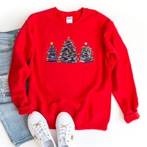 A red sweater with a Christmas tree design, a pair of blue jeans, and white sneakers are laid out on a white background.