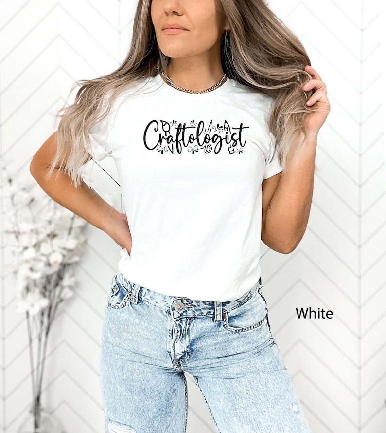 May include: A white t-shirt featuring the text "Craftologist" in black lettering with decorative floral accents. The shirt is being worn by a person with long blonde hair and blue jeans.
