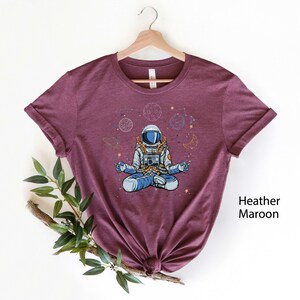 May include: A heather maroon t-shirt featuring a cartoon illustration of an astronaut meditating in space. The astronaut is surrounded by planets, stars, and crescent moons.