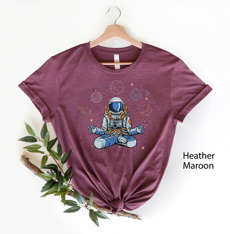May include: A heather maroon t-shirt featuring a cartoon illustration of an astronaut meditating in space. The astronaut is surrounded by planets, stars, and crescent moons.