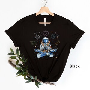 May include: Black t-shirt with a graphic of an astronaut meditating in space with planets and stars. The text "Black" is visible in the bottom right corner of the image.