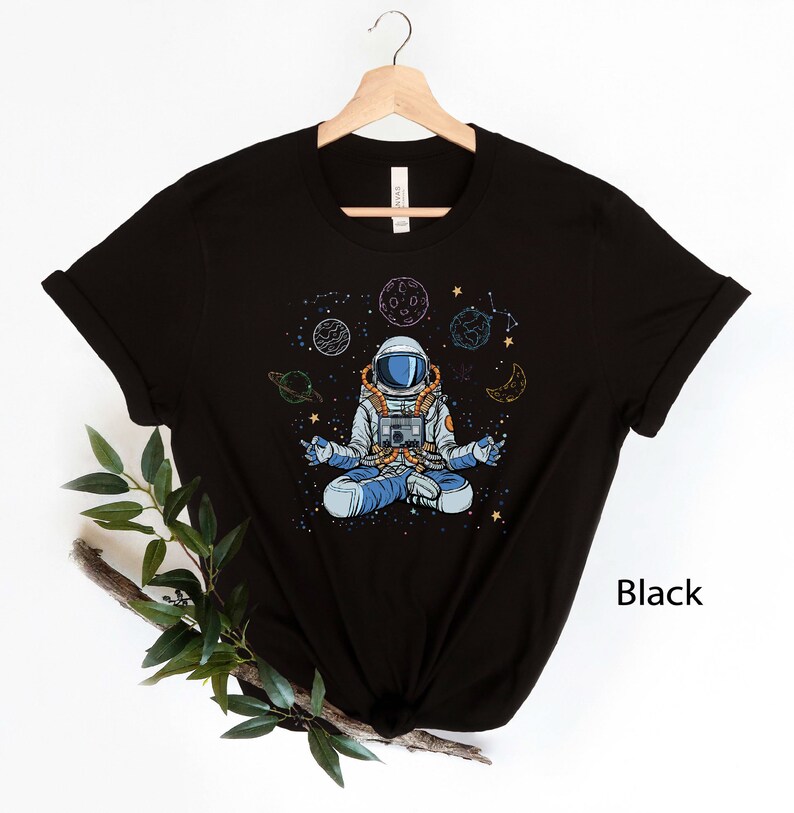May include: Black t-shirt with a graphic of an astronaut meditating in space with planets and stars. The text "Black" is visible in the bottom right corner of the image.