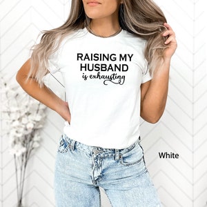 May include: White t-shirt with black text that reads "Raising my husband is exhausting".