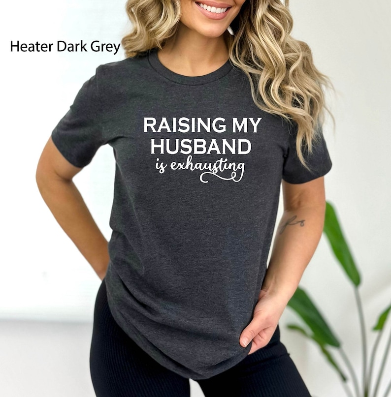 May include: A dark grey t-shirt with the text "Raising my husband is exhausting" in white.
