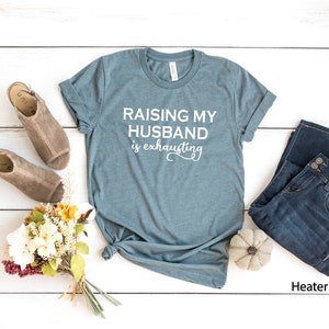 May include: A teal blue t-shirt with the text "Raising my husband is exhausting" in white lettering. The shirt is laid flat on a white wooden surface with a pair of brown sandals, a bouquet of flowers, a pair of blue jeans, and a small white pumpkin.