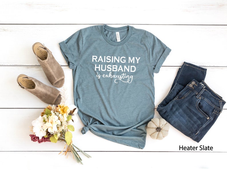 May include: A teal blue t-shirt with the text "Raising my husband is exhausting" in white lettering. The shirt is laid flat on a white wooden surface with a pair of brown sandals, a bouquet of flowers, a pair of blue jeans, and a small white pumpkin.