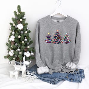 A gray sweatshirt with a Christmas tree design on it, along with a small Christmas tree, a pair of jeans, and some ornaments, all set against a white background.
