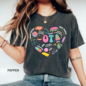 May include: A dark gray t-shirt with a colorful graphic design of a heart shape with the word "LOT" in the center. The heart is surrounded by various images of toys and activities, including a walker, a swing set, a ball, a puzzle, and a paint palette. The words "visual motor", "fine motor", "social", "cognition", "sensory processing", "play", "bilateral coordination", and "executive functioning" are written around the heart.