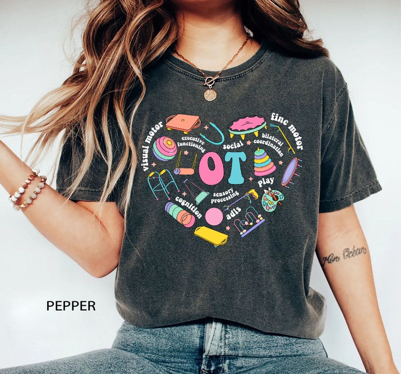 May include: A dark gray t-shirt with a colorful graphic design of a heart shape with the word "LOT" in the center. The heart is surrounded by various images of toys and activities, including a walker, a swing set, a ball, a puzzle, and a paint palette. The words "visual motor", "fine motor", "social", "cognition", "sensory processing", "play", "bilateral coordination", and "executive functioning" are written around the heart.