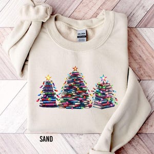 A beige sweater with a graphic design of three Christmas trees made out of books, each tree having a star on top.