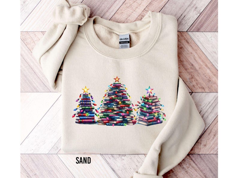 A beige sweater with a graphic design of three Christmas trees made out of books, each tree having a star on top.