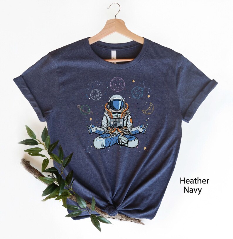 May include: A navy blue t-shirt with a graphic of an astronaut meditating in space. The astronaut is surrounded by planets, stars, and a crescent moon. The text "Heather Navy" is visible in the bottom right corner of the image.