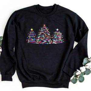 A black sweatshirt with a graphic design of three Christmas trees made of stacked books, surrounded by white cotton balls and greenery.