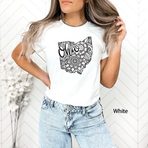 May include: A white t-shirt with a black outline of the state of Ohio filled with a mandala design. The word "Ohio" is written in black inside the state outline. The t-shirt is being worn by a person with long brown hair. The background is a white wall with a chevron pattern.