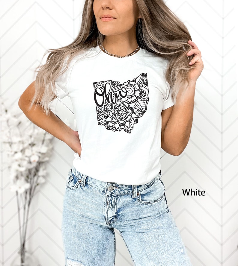 May include: A white t-shirt with a black outline of the state of Ohio filled with a mandala design. The word "Ohio" is written in black inside the state outline. The t-shirt is being worn by a person with long brown hair. The background is a white wall with a chevron pattern.