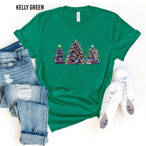 A green t-shirt with a Christmas tree design, a pair of blue jeans, and a pair of white sneakers are laid out on a white background.