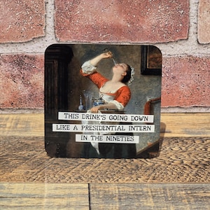 May include: Square coaster with a vintage painting of a woman drinking. The coaster has the text "THIS DRINK'S GOING DOWN LIKE A PRESIDENTIAL INTERN IN THE NINETIES" in white font. The background is a brick wall and wooden surface.