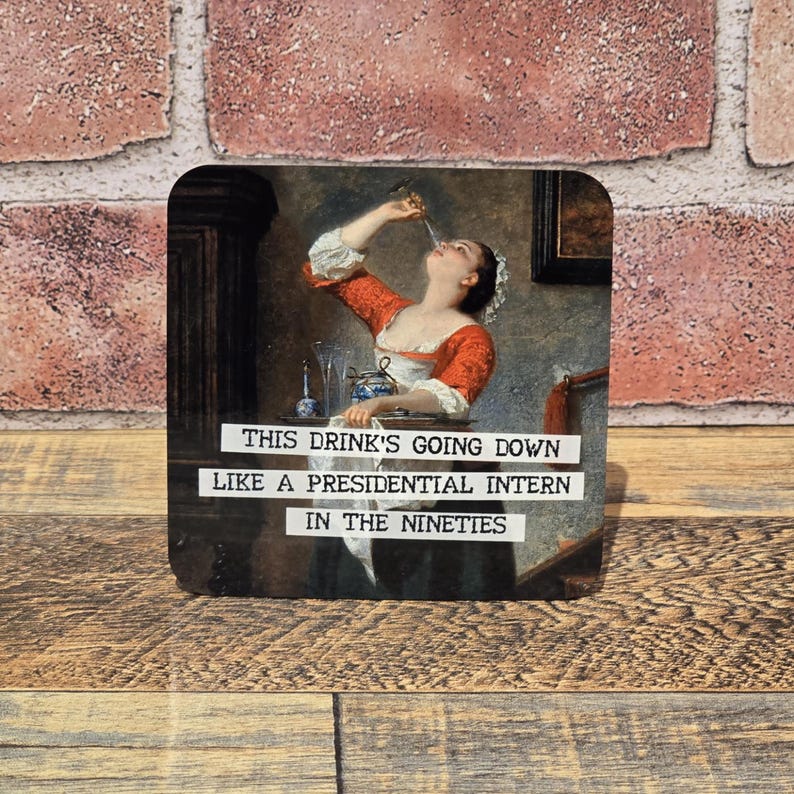 May include: Square coaster with a vintage painting of a woman drinking. The coaster has the text "THIS DRINK'S GOING DOWN LIKE A PRESIDENTIAL INTERN IN THE NINETIES" in white font. The background is a brick wall and wooden surface.