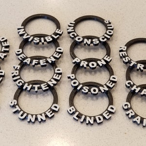 May include: A set of 18 black plastic rings with white lettering. Each ring has a different word printed on it, such as "Invisible", "Deafened", "Prone", "Restrained", "Charmed", "Grappled", "Incapacitated", "Paralyzed", "Petrified", "Stunned", "Blinded", and "Poisoned".
