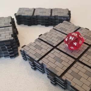 Peut inclure: Trois piles de dalles de jeu grises &agrave; motif de briques avec des bords noirs. Un d&eacute; rouge &agrave; vingt faces repose sur une pile. Les dalles sont con&ccedil;ues pour les jeux de table, cr&eacute;ant un environnement de donjon ou de b&acirc;timent.