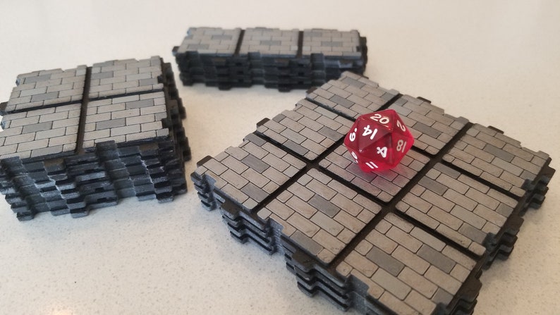 Peut inclure: Trois piles de dalles de jeu grises &agrave; motif de briques avec des bords noirs. Un d&eacute; rouge &agrave; vingt faces repose sur une pile. Les dalles sont con&ccedil;ues pour les jeux de table, cr&eacute;ant un environnement de donjon ou de b&acirc;timent.