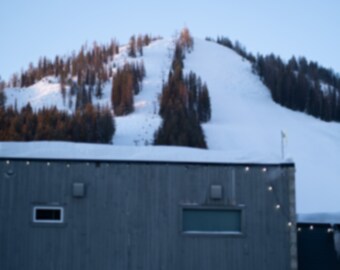 Pode incluir: Um edifício de metal cinza com três janelas e um telhado coberto de neve. O edifício está em frente a uma montanha coberta de neve com árvores.