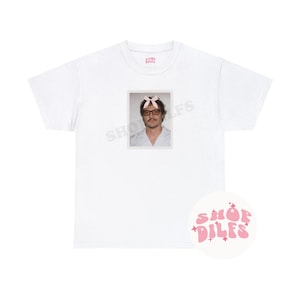 May include: White t-shirt featuring a framed image of a person wearing glasses and a pink bow. The shirt also has the text "SHOP DILFS" in a pink, retro font on a white circle.