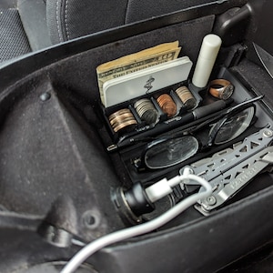 May include: A black car center console organizer with a variety of items, including cash, coins, a pen, glasses, a multi-tool, and a phone charger.
