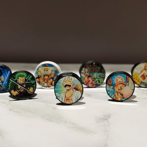 以下が含まれることがあります： カラフルなアニメキャラクターのデザインが施された、丸い白と黒のポップソケットのコレクション。ポップソケットには、海賊をテーマにしたものなど、さまざまなキャラクターイラストが描かれており、大理石の表面に表示されています。