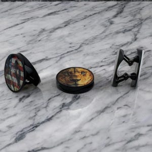 Op de afbeelding: Drie telefoonbeugels met verschillende ontwerpen. De eerste beugel heeft een Amerikaans vlagontwerp. De tweede beugel heeft een leeuwenkopontwerp. De derde beugel is een eenvoudig zwart ontwerp.