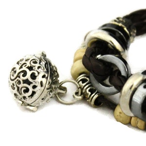 May include: A brown leather bracelet with silver and white beads, a silver charm with a decorative design, and a silver bell charm.