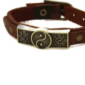 May include: A brown leather bracelet with a silver-toned metal charm featuring a yin yang symbol and the Bagua, an eight trigram symbol.