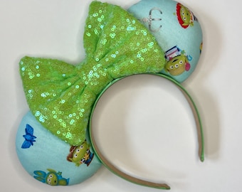 Puede incluir: Diadema con lazo verde de lentejuelas y orejas de tela azul claro con personajes de Toy Story, incluyendo Woody, Buzz Lightyear y Alien.