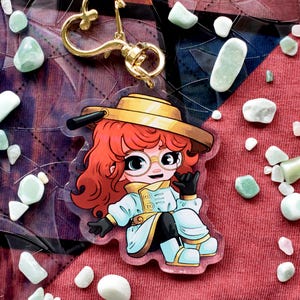 May include: A clear acrylic charm featuring a cartoon character with red hair, glasses, and a yellow hat. The charm has a gold-colored heart-shaped clasp. The background is a red fabric with scattered white and green stones.