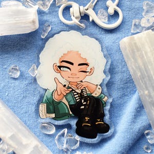 Puede incluir: Un dije acrílico transparente con un personaje de dibujos animados con cabello rizado blanco, una chaqueta verde y botas negras. El dije tiene un cierre blanco y está rodeado de cristales transparentes sobre un fondo de tela azul.
