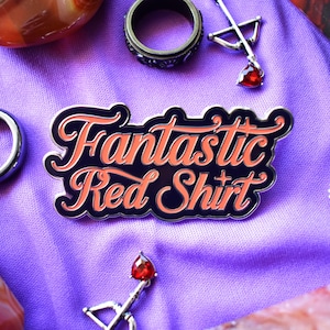 Puede incluir: Un pin de esmalte negro y rojo con el texto "Fantastic Red Shirt" en una fuente estilizada. El pin está rodeado de joyas, incluyendo un anillo y un colgante con forma de arco y flecha con una piedra preciosa roja.