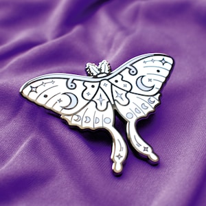 Può includere: Una spilla a forma di falena in smalto color argento con dettagli bianchi e neri. La spilla presenta lune crescenti, stelle e altri disegni celesti. La spilla è posta su uno sfondo di tessuto viola.