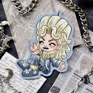 Puede incluir: Una pegatina con un personaje de dibujos animados con cabello rubio rizado, vistiendo un atuendo gris y señalando. La pegatina está sobre un fondo gris con cadenas y recortes de periódicos.