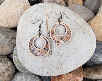 May include: A pair of wooden hoop earrings with a floral design. The earrings feature a double-hoop design with a clear center, showcasing a pattern of orange and purple flowers and leaves. The metal ear wires are a dark bronze color.