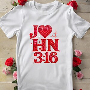 Puede incluir: Camiseta blanca con un corazón rojo y el texto "JOHN 3:16" en rojo. El diseño presenta un gráfico en forma de corazón y acentos brillantes. La camiseta está rodeada de rosas rojas y rosas.