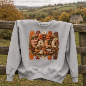 May include: A light grey crewneck sweatshirt featuring a central graphic with the word FALL in large, textured letters. The design includes an orange bow, pumpkins, autumn leaves, and floral elements arranged in a wreath-like pattern.