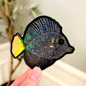 May include: A black and iridescent fish sticker with a yellow fin. The fish has a speckled pattern and a round body.