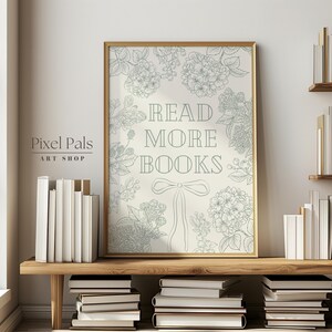 May include: A framed print with the text "READ MORE BOOKS" in a decorative font, surrounded by floral illustrations. The print is in a gold frame and is displayed on a wooden shelf with stacks of books. The overall aesthetic is a cosy, literary theme.