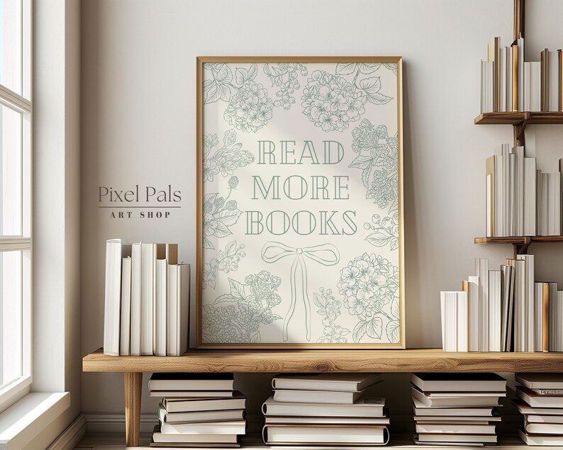 May include: A framed print with the text "READ MORE BOOKS" in a decorative font, surrounded by floral illustrations. The print is in a gold frame and is displayed on a wooden shelf with stacks of books. The overall aesthetic is a cosy, literary theme.
