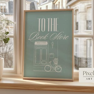 May include: A framed art print with the words "TO THE Book Store" in white script on a teal background. The illustration features a bicycle, a storefront, and a street lamp. The print is displayed on a wooden windowsill.
