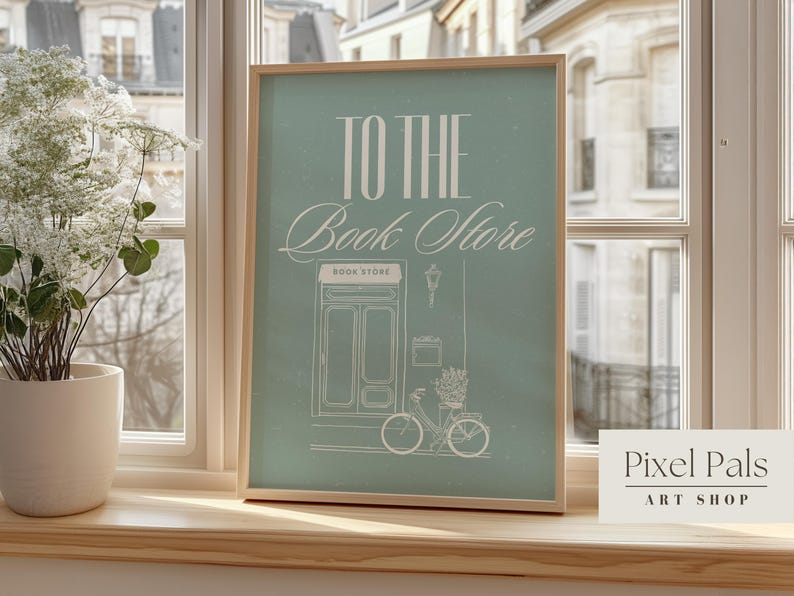 May include: A framed art print with the words "TO THE Book Store" in white script on a teal background. The illustration features a bicycle, a storefront, and a street lamp. The print is displayed on a wooden windowsill.