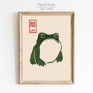 May include: Framed art print featuring a green frog illustration with a large open mouth. The frog is set against a cream background, with a red Japanese character in the upper left corner. The print is in a wooden frame.