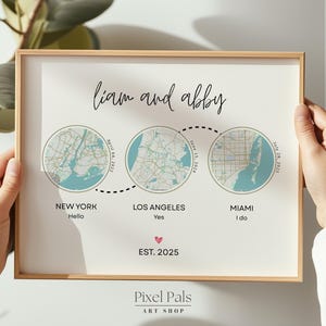May include: A framed print with three circular map illustrations linked by a dotted line, representing New York, Los Angeles, and Miami. The print includes the names "Liam and Abby" and the text "EST. 2025". The print is held by hands.