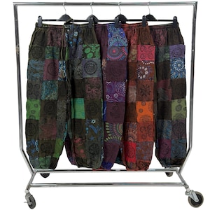 Op de afbeelding: Een kledingrek toont vijf patchwork broeken. De broeken hebben verschillende kleuren, waaronder groen, blauw, paars en rood. Elke broek heeft een uniek patroon en is gemaakt van een zachte, vloeiende stof. De broeken hangen aan zwarte hangers.